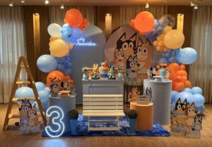 Decoração infantil tema Bluey para locação, com painel personalizado, personagens, mesa decorativa e arco orgânico de balões em azul, laranja, branco e amarelo.