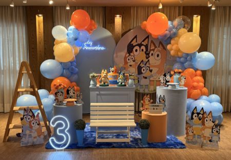 Decoração infantil tema Bluey para locação, com painel personalizado, personagens, mesa decorativa e arco orgânico de balões em azul, laranja, branco e amarelo.